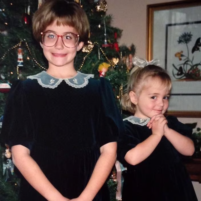 Paige and her younger sister, Holly, pose in front of a Christmas tree wearing matching black velvet dresses with lace Peter Pan collars.